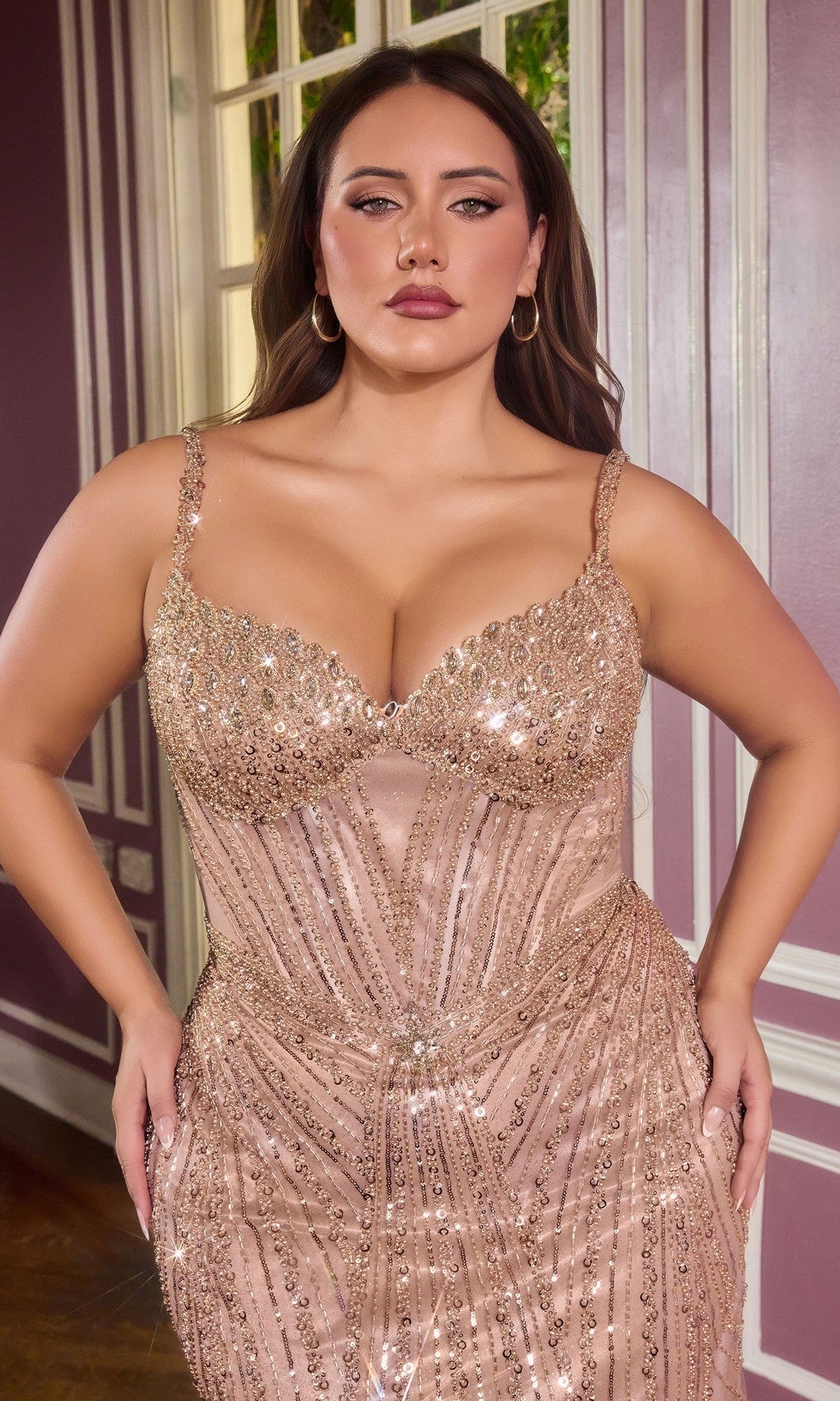A woman with long brown hair stands indoors by a window, wearing the sparkling, form-fitting Ladivine CC398C Long Plus-Size Prom Dress against mauve walls with white trim.